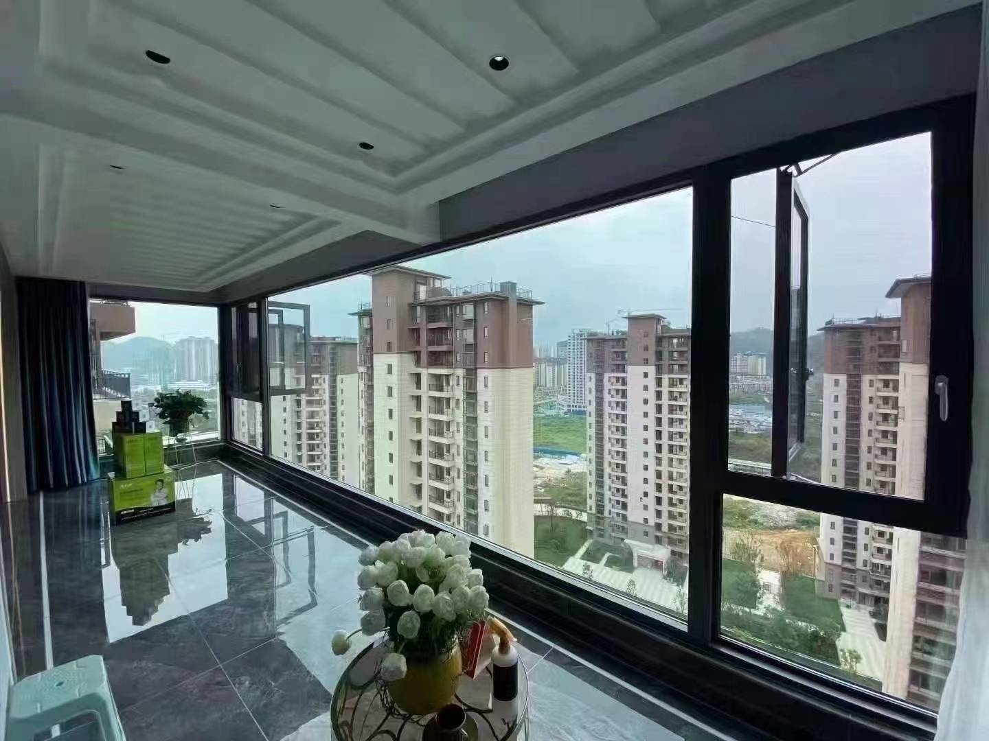高层住宅阳台封窗实景,大面积黑色断桥铝落地窗占据主要视野,部分窗扇开启,窗外可见密集的城市楼盘与远山景观。室内铺设光面灰色大理石纹地砖,倒映出窗外景色,天花板采