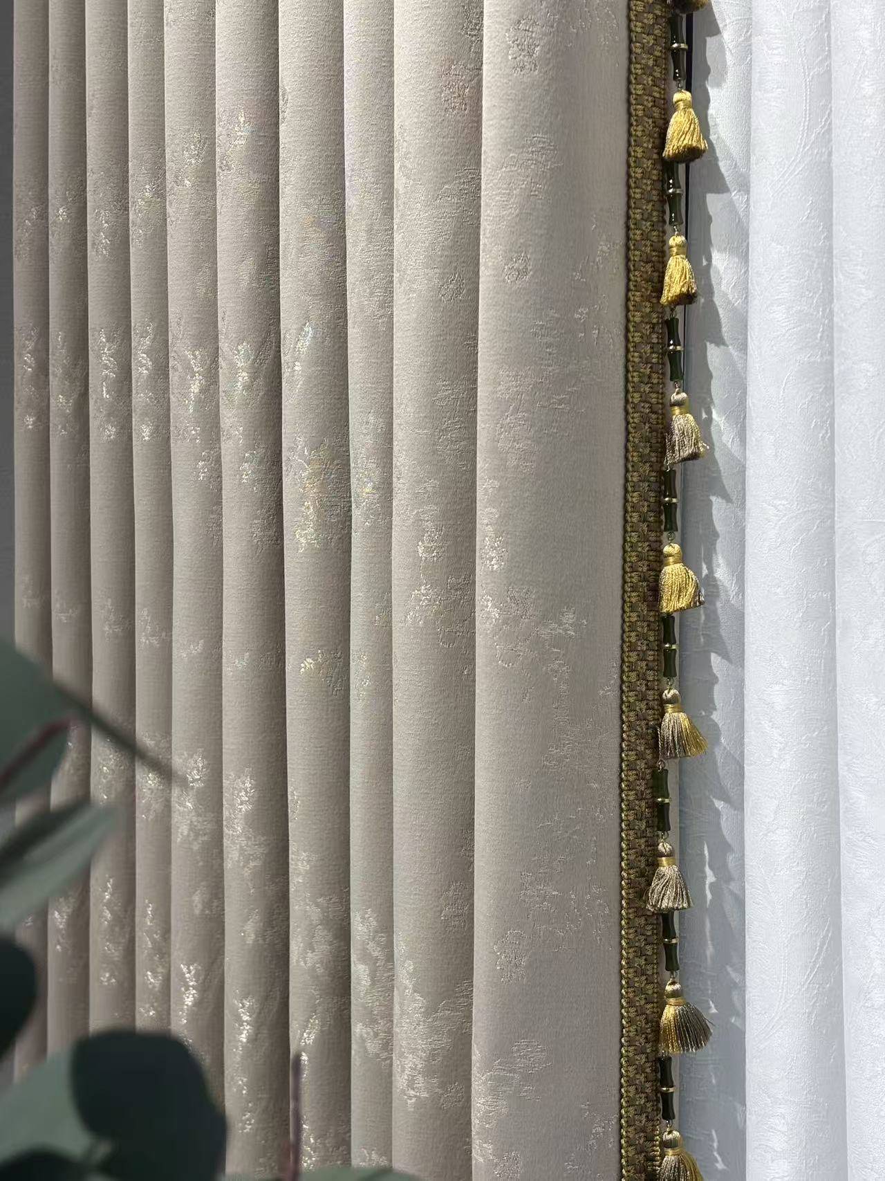 这是一张展示浅灰色高精密提花窗帘的特写图片。窗帘面料带有细腻的暗纹图案，质感厚实且垂坠感极佳，边缘装饰有精致的金色流苏花边，增添了轻奢氛围。右侧搭配白色半透明纱