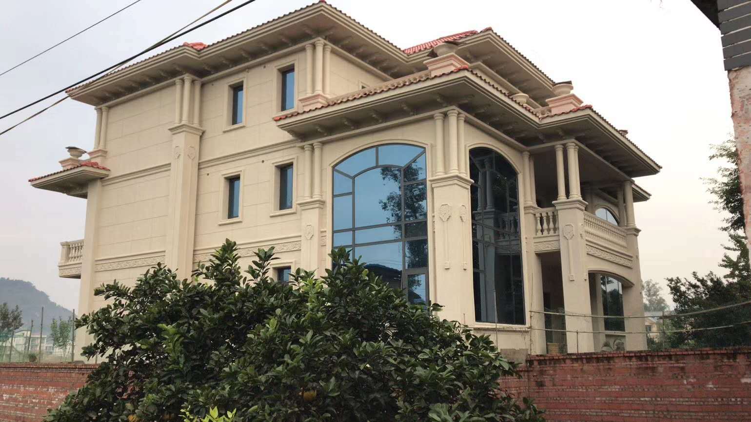 这是一栋位于乡村环境的米黄色欧式独栋别墅。建筑采用米黄色外墙搭配红褐色坡屋顶，设有大型拱形落地窗和罗马柱装饰，采光良好且外观大气。墙面带有精致的浮雕线条和阳台栏