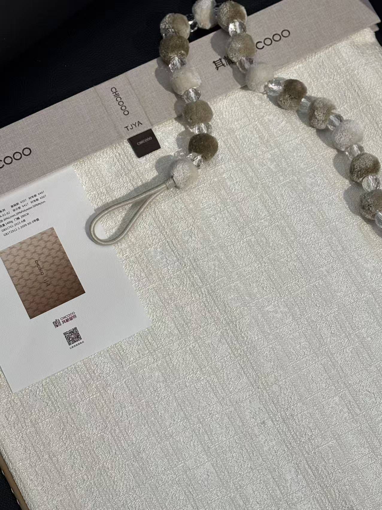 这是一张展示CHICOOO其趣窗帘面料样册的特写图片。画面主体为一块白色肌理感窗帘布，表面呈现细腻的竖条纹雪尼尔质感，搭配一串由灰绿色毛绒球和透明水晶珠交替串成