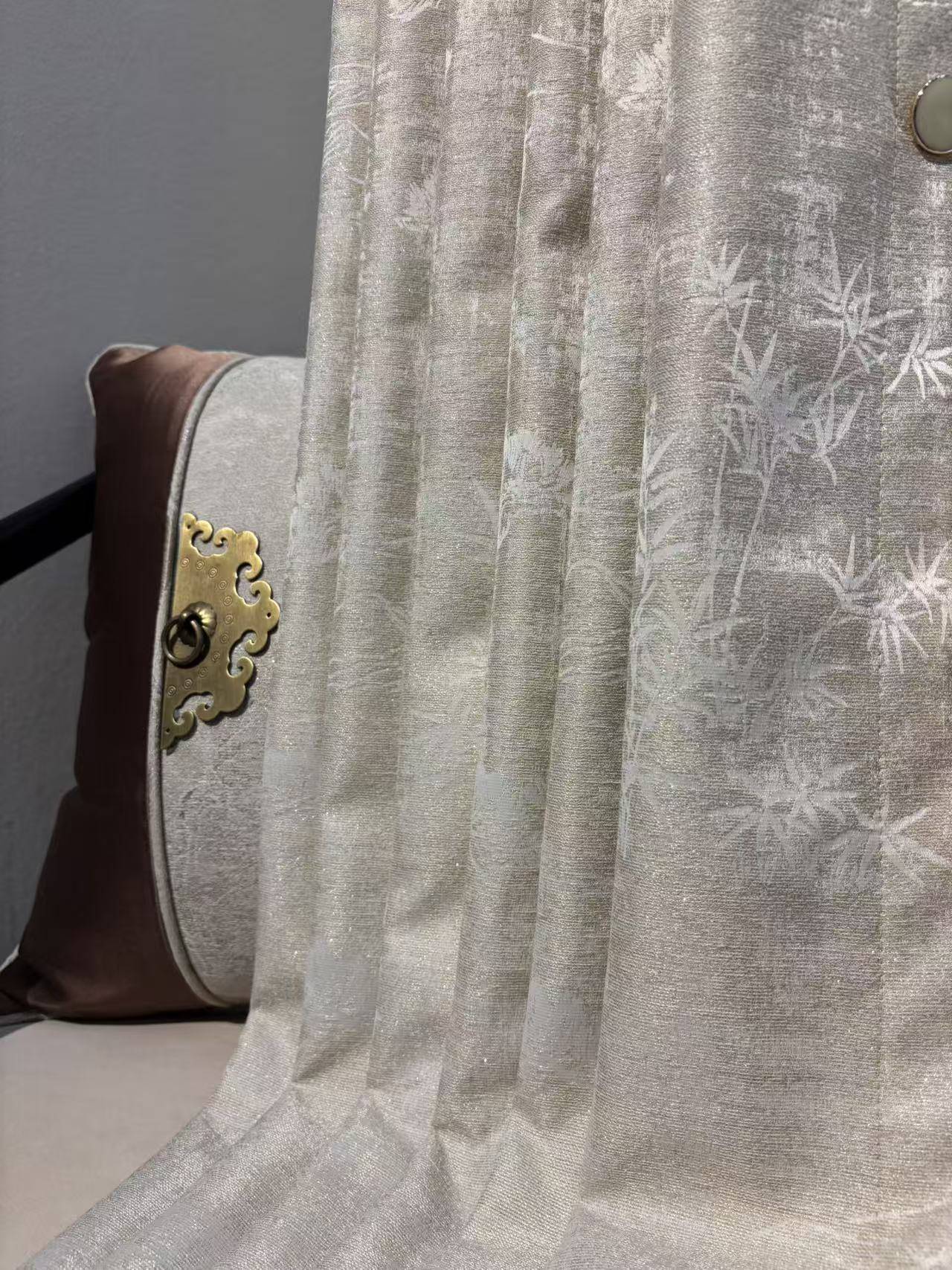 居家样板间一角，展示了一款银灰色新中式提花窗帘。面料带有细腻的珠光光泽，织有淡雅的竹叶暗纹，垂坠自然。左侧搭配深棕色抱枕，点缀金色云纹金属环扣，增添古典雅致气息