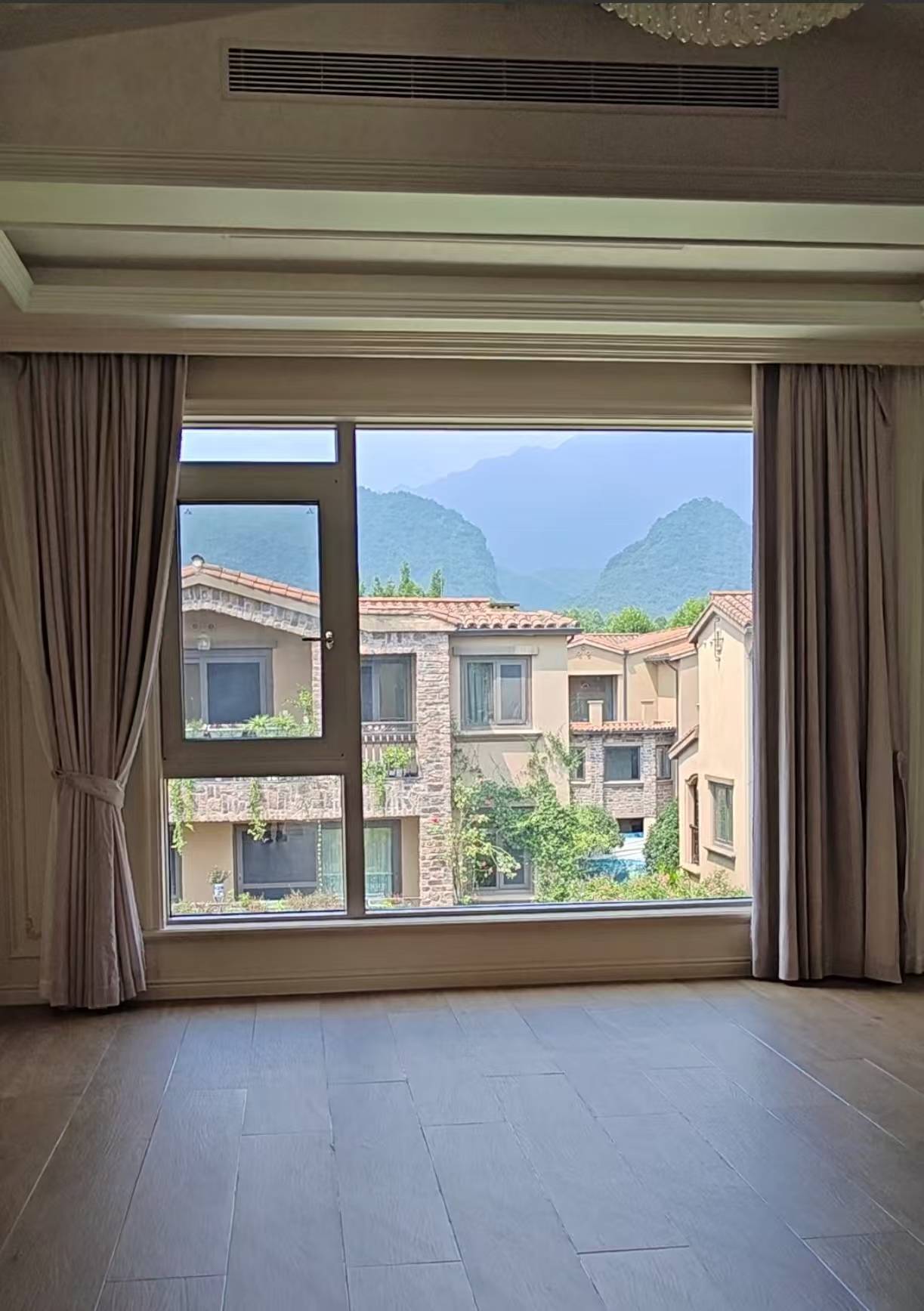 这是一张展示别墅室内景观的图片。画面主体为一扇宽大的落地窗，采用深灰色窗框，左侧设有开启扇，保证通风采光。两侧搭配米灰色落地窗帘，质感垂顺。室内铺设浅灰色木地板