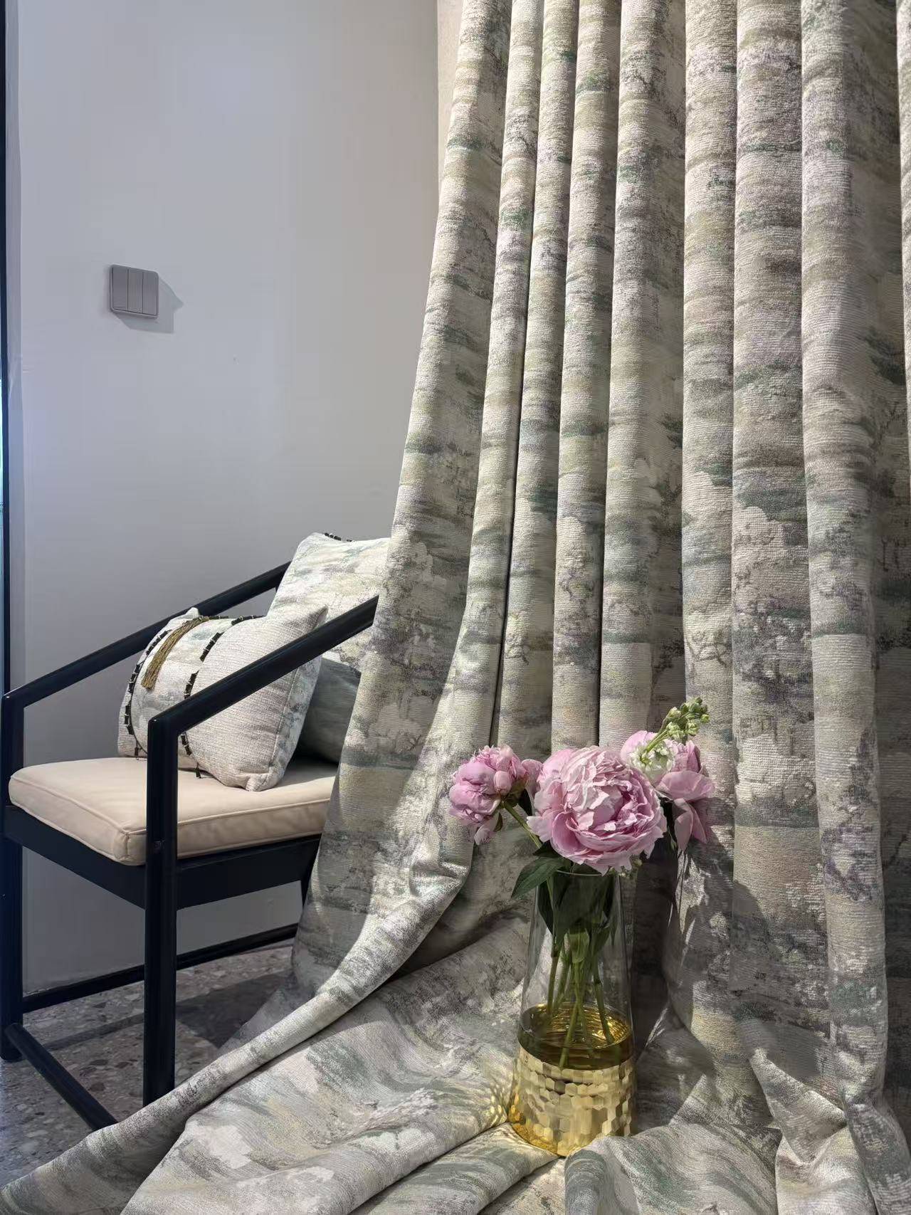 这是一张展示新中式家居软装搭配的图片。画面主体为灰绿色调的落地窗帘，面料带有抽象的水墨山水纹理，垂坠感极佳，营造出雅致的东方韵味。左侧搭配一把黑色金属框架的新中