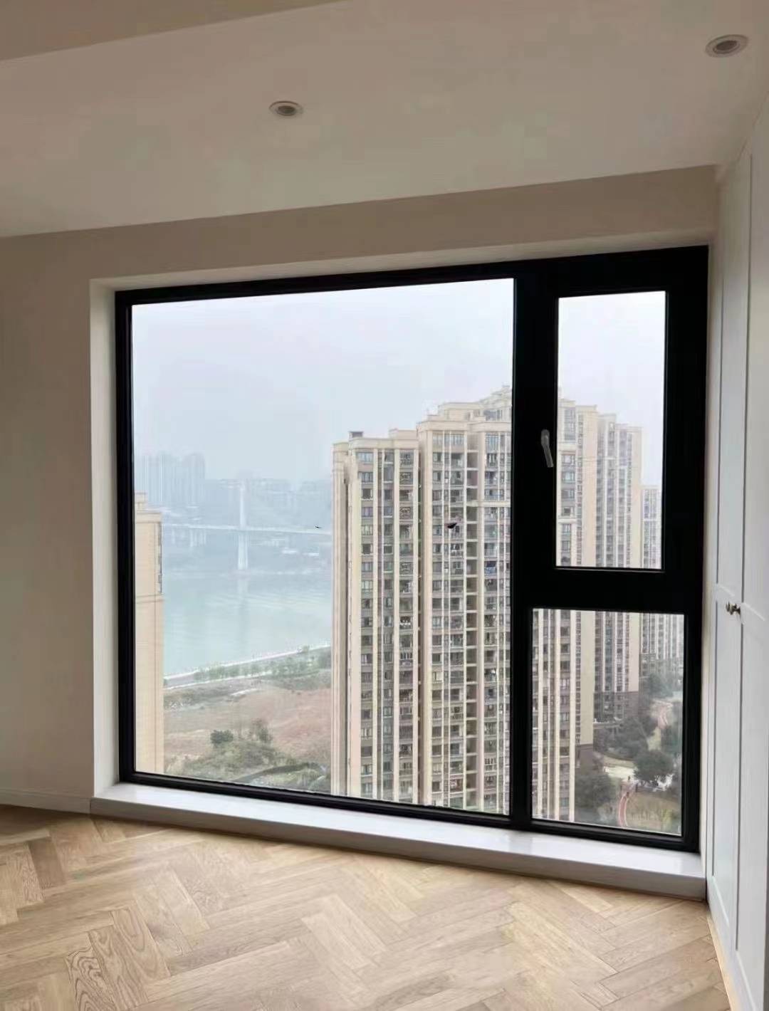 高层住宅室内实景，安装黑色边框大面积落地窗，右侧配置上下分格开启扇。搭配浅色人字拼木地板与白色石材窗台，现代简约风格。窗外直面江景与城市高楼，采光通透，视野开阔