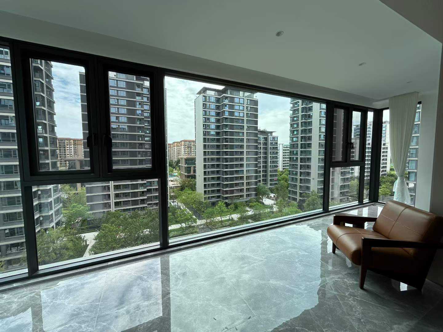 高层住宅阳台封窗实景，采用黑色断桥铝系统窗，大面积落地玻璃搭配两侧开启扇，视野开阔无遮挡。室内铺设灰色大理石纹亮面地砖，倒影清晰，搭配棕色皮质沙发与白色纱帘，营