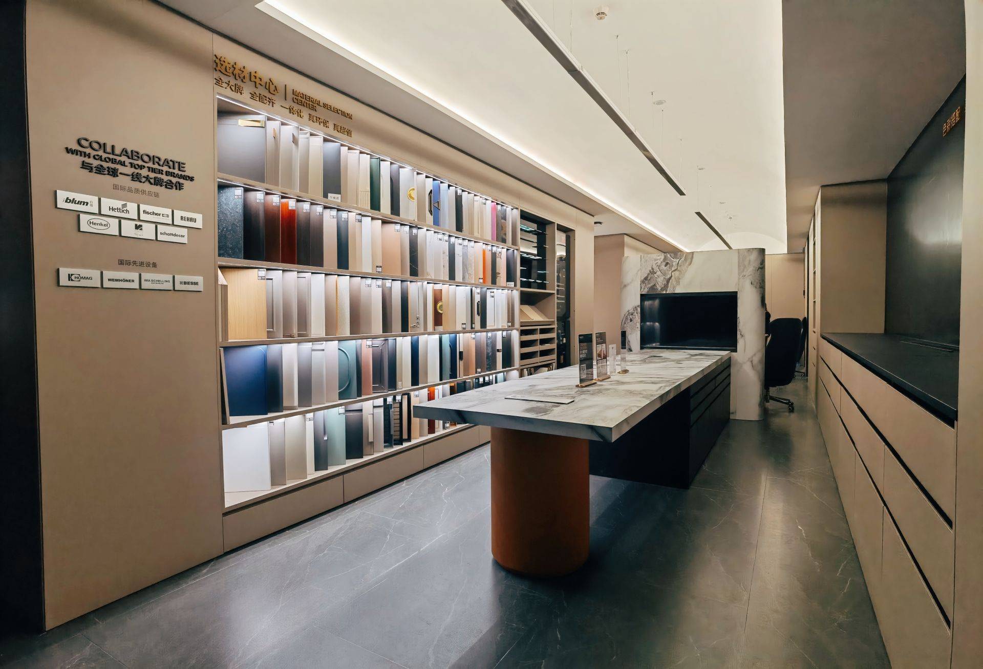 高端家居建材展厅选材中心实景，墙面整齐陈列多色板材样品与木纹饰面，中央摆放岩板台面岛台搭配深色圆柱底座，地面铺设深灰色纹理地砖，整体现代简约风格，展示国际一线品