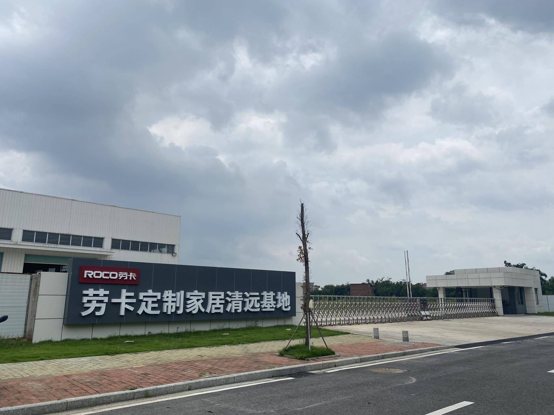 图片展示了劳卡定制家居清远基地的入口实景。左侧矗立着深灰色大型招牌，上方有红色“ROCO劳卡”标识，下方为白色立体大字“劳卡定制家居清远基地”。背景可见白色现代