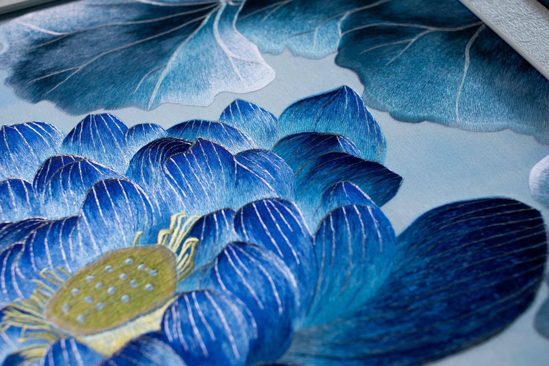 这是一幅精美的新中式风格刺绣装饰画特写。画面主体为盛开的蓝色莲花与舒展的荷叶，采用细腻的苏绣工艺，丝线呈现出丰富的光泽感与立体层次。花瓣由深蓝至浅蓝自然渐变，搭