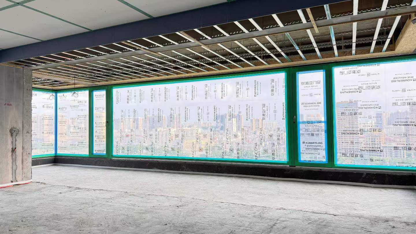 这是一处正在施工的室内场景，天花板裸露轻钢龙骨，地面为水泥基层。画面主体为一排新安装的落地系统门窗，玻璃表面覆盖着印有“京港亚门窗”、“KOMMERLING”柯