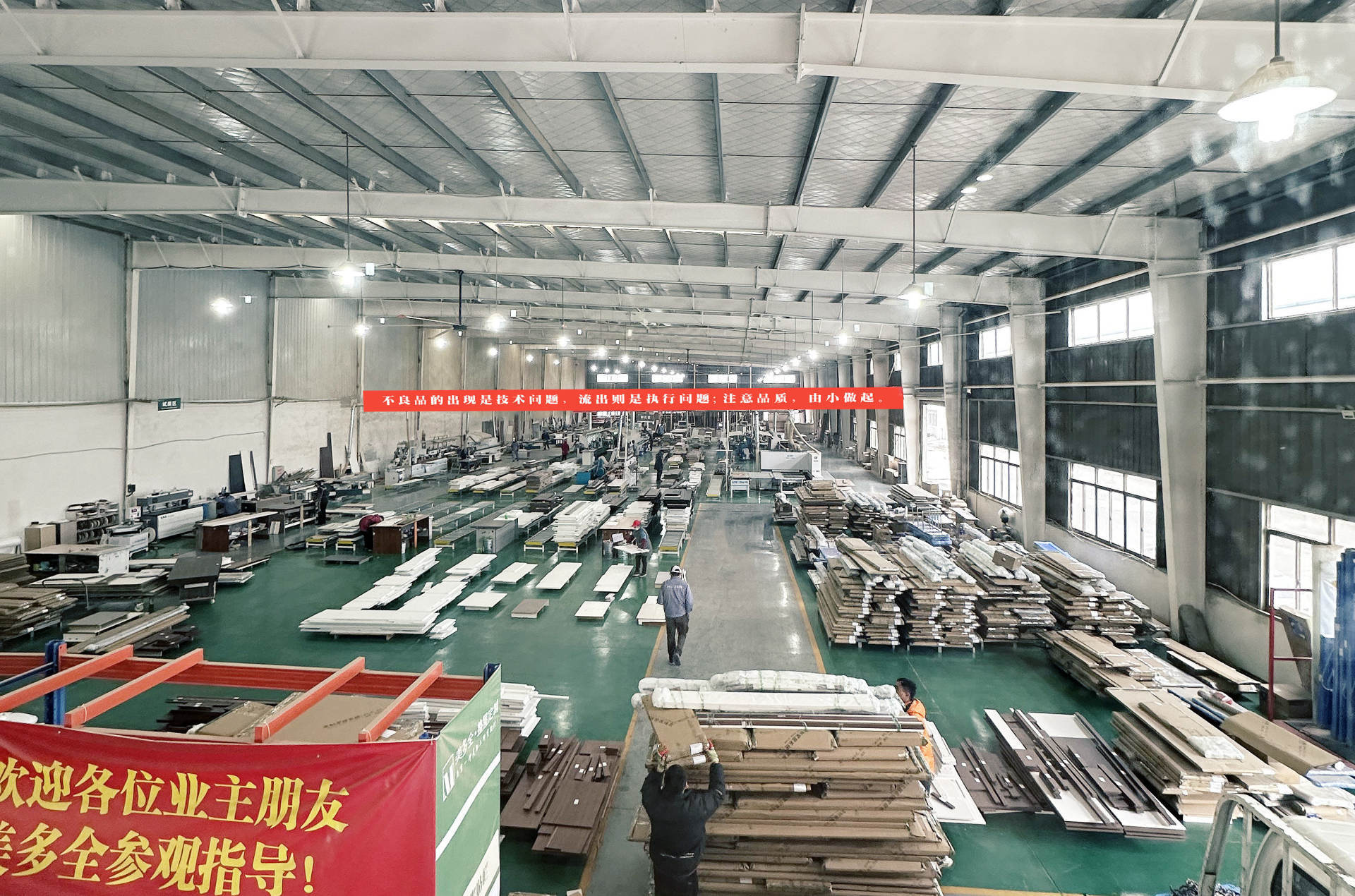 大型家具工厂车间全景，绿色环氧地坪，钢结构厂房顶部。车间内整齐堆放大量板材原料和半成品，配备专业加工机械设备。工人正在搬运和加工板材，红色横幅悬挂于车间中央，强