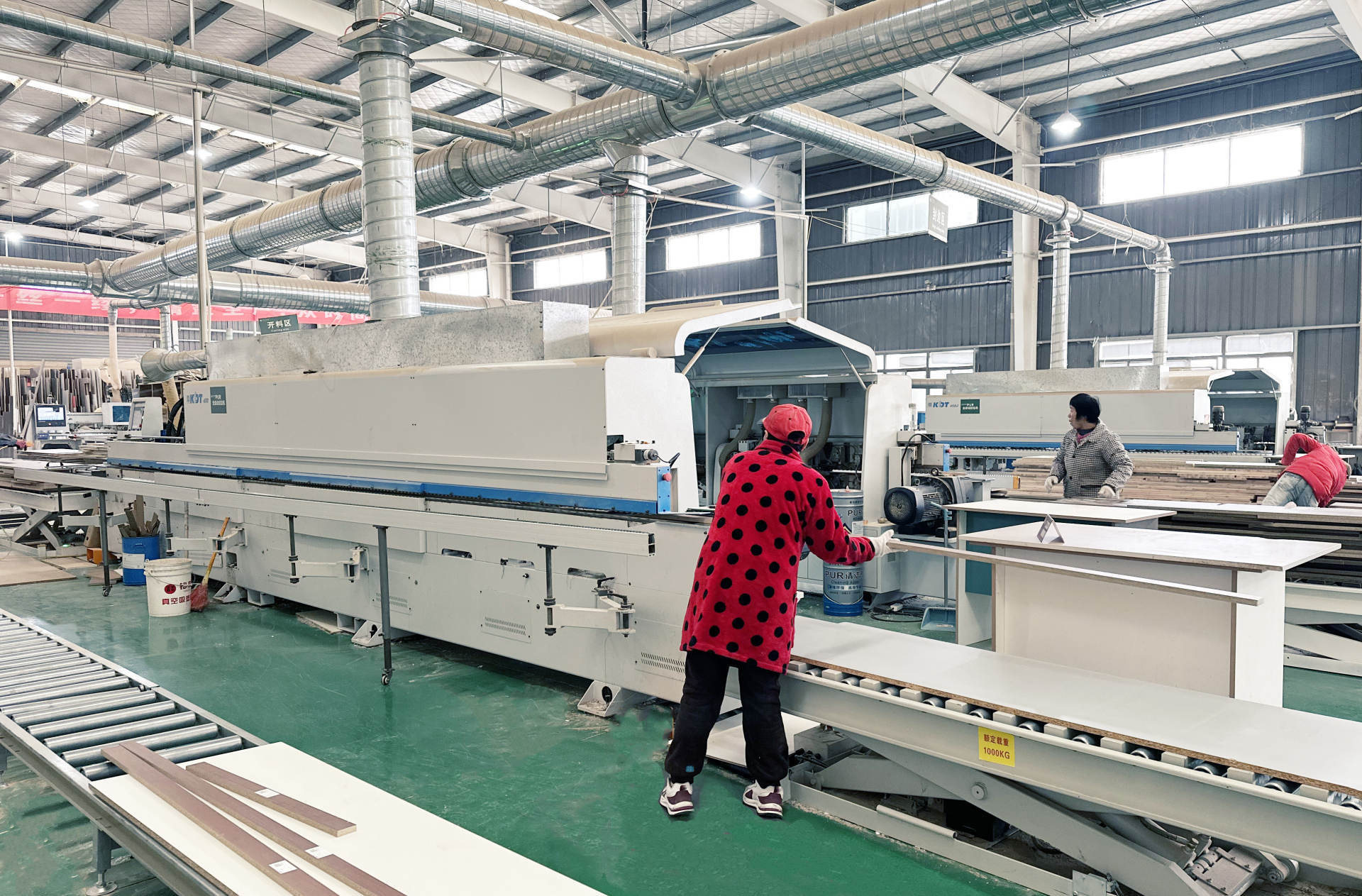 宽敞明亮的木工制造工厂车间内，大型白色KDT品牌木工机械正在运行，顶部布满银色通风管道。前景身穿红色波点外套的工人正在操作传送带上的白色板材，机器旁放置蓝色PU